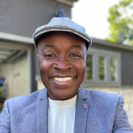 Headshot of Stephen Quaye smiling and wearing a plaid hat and blue-gray suit
