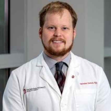 Headshot of Nicholas Denton wearing a white lab coat and gray and red tie.